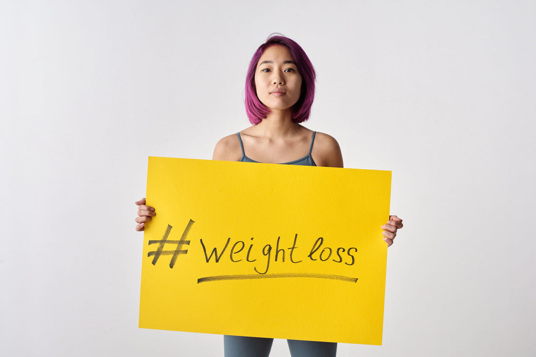 Asian woman with purple hair holding a weight loss sign in a studio setting.